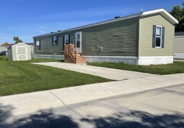 A light green manufactured home with white trim, a small wooden staircase leading to the door, a shed in the grassy yard, and a concrete driveway on a sunny day.