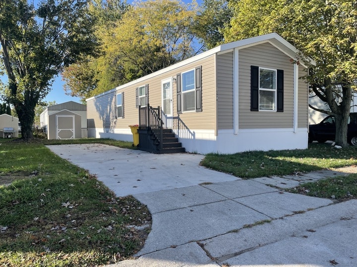 A beige mobile home with white trim, black shutters, and a small front porch sits next to a concrete driveway. There is a yellow trash bin and a matching storage shed in the grassy yard, with trees and blue sky in the background.