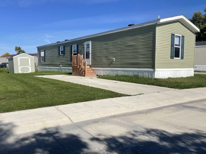 A light green manufactured home with white trim, a small wooden staircase leading to the door, a shed in the grassy yard, and a concrete driveway on a sunny day.