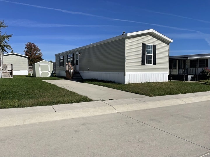A beige manufactured home with white trim sits on a grassy lot with a small driveway and front steps. A shed is in the backyard, and another similar home is visible next door under a clear blue sky.