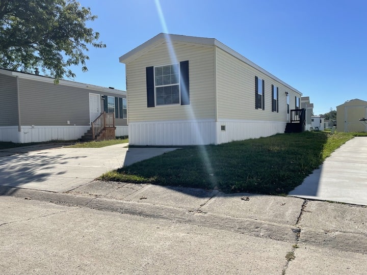 A beige manufactured home with black shutters sits on a grassy lot beside a paved driveway under a clear blue sky. Another similar home is visible to the left. Sunlight casts a bright beam across the scene.