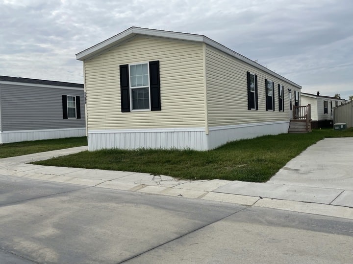 A beige manufactured home with black shutters sits on a grassy lot beside a concrete driveway. Another similar home is visible to the left under a cloudy sky.
