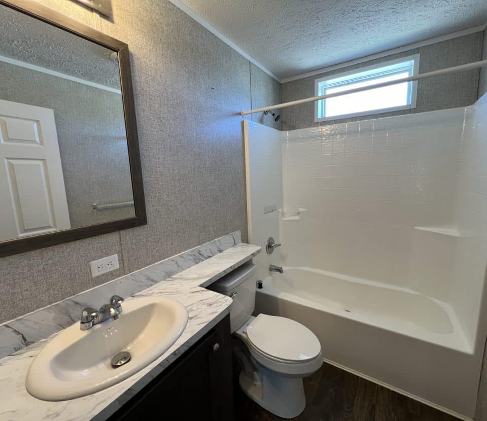 Modern bathroom with a single sink, marble countertop, oval mirror, toilet, and a white bathtub-shower combo with a window above. The walls have a textured finish and the floor is wood-style.