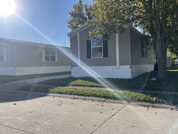 A beige manufactured home with white trim sits in the sunlight near a tree, casting a shadow on the grass. A sunbeam stretches diagonally across the image, and another similar home is visible next door.