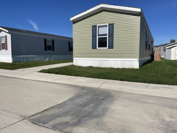 A light green manufactured home with blue shutters sits on a grassy lot beside a paved driveway. Another similar home is visible to the left. The sky is clear and blue.