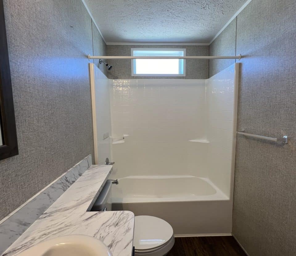 A small bathroom with a bathtub and shower, a white toilet, a marble-patterned countertop with a sink, a towel bar on the right wall, and a window above the tub letting in natural light.