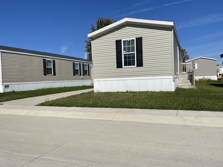 A beige manufactured home with white trim and black shutters sits beside a driveway and patch of green grass under a clear blue sky, with another similar home nearby.