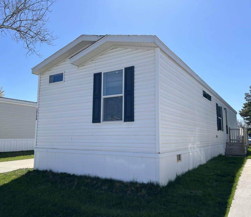 A white mobile home with blue shutters sits on a grassy lot under a clear blue sky, with a driveway and a white SUV parked nearby.