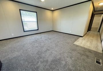 A spacious, empty room with beige walls, gray carpet, a large window, and a section of light wood flooring near a hallway leading to another area. A ceiling fan with a light is visible above.