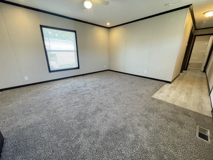 A spacious, empty room with beige walls, gray carpet, a large window, and a section of light wood flooring near a hallway leading to another area. A ceiling fan with a light is visible above.