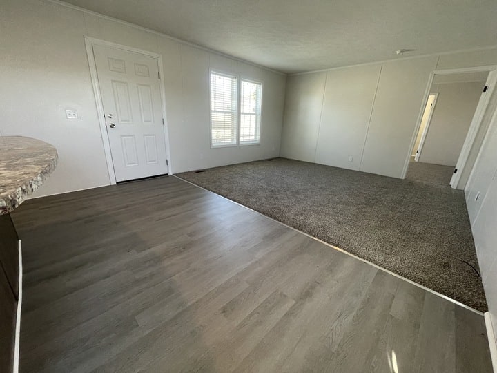 A bright, empty room with light gray wood flooring near the entrance, transitioning to carpet in the living area. White walls, a front door, two large windows, and a doorway to another room are visible.