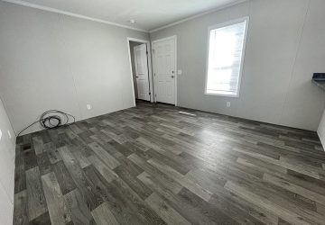 Empty room with gray wood-style flooring, light gray walls, a white door, and a window letting in natural light. Coiled cables are on the floor near one wall. The space appears clean and unfurnished.