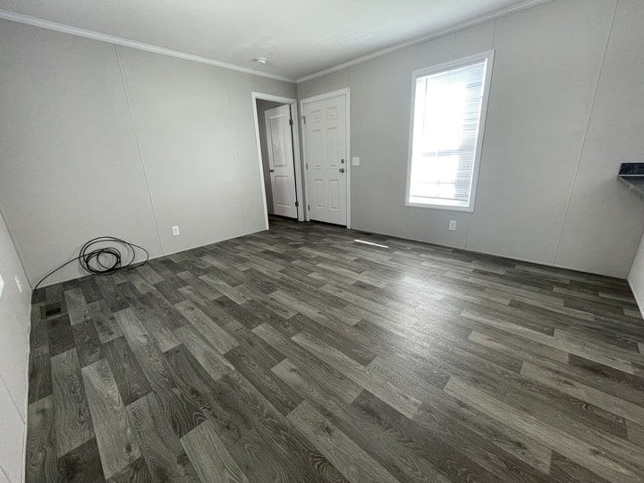 Empty room with gray wood-style flooring, light gray walls, a white door, and a window letting in natural light. Coiled cables are on the floor near one wall. The space appears clean and unfurnished.