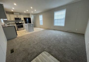 Open-concept living area with gray carpet, large window, and adjoining modern kitchen featuring stainless steel appliances, white cabinetry, and an island with bar seating. Light wood-look flooring in the kitchen.