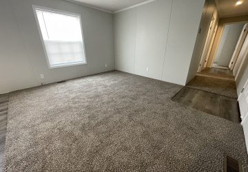 Empty room with light gray carpet and plain light-colored walls, a large window with blinds on the left, and a hallway with doorways visible on the right. One corner has a small section of wood flooring.