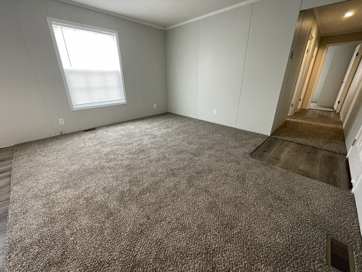 Empty room with light gray carpet and plain light-colored walls, a large window with blinds on the left, and a hallway with doorways visible on the right. One corner has a small section of wood flooring.