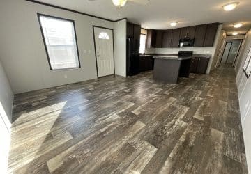 Open concept kitchen and living area with wood-look flooring, brown cabinets, center island, neutral walls, windows, a ceiling fan, and a door leading outside. Room is empty and brightly lit by natural light.