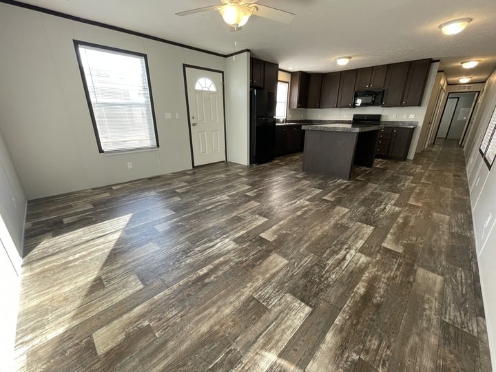 Open concept kitchen and living area with wood-look flooring, brown cabinets, center island, neutral walls, windows, a ceiling fan, and a door leading outside. Room is empty and brightly lit by natural light.