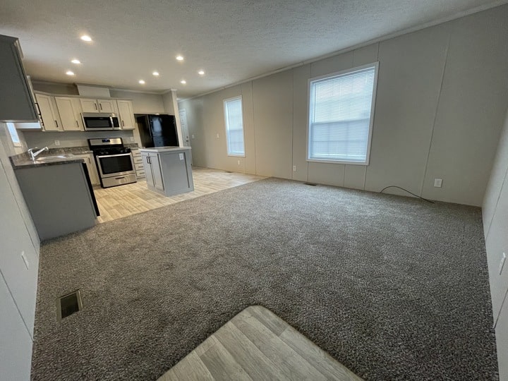 Open-concept living area with gray carpet, large window, and adjoining modern kitchen featuring stainless steel appliances, white cabinetry, and an island with bar seating. Light wood-look flooring in the kitchen.