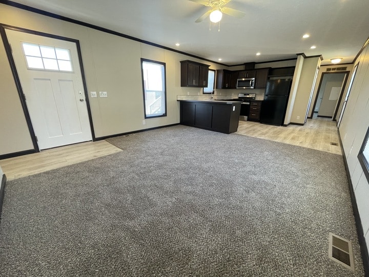 Open living area with gray carpet and beige walls, adjacent to a modern kitchen with dark cabinets, stainless steel appliances, and light wood flooring; front door and windows provide ample natural light.