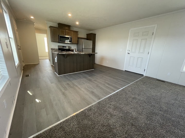 Open-concept room with light wood flooring and an adjoining carpeted area. Modern kitchen features dark wood cabinets, stainless steel appliances, and an island. White walls and a door are visible on the right.