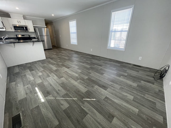 A modern, unfurnished room with gray wood-look flooring, white walls, two large windows, and a kitchen area featuring stainless steel appliances and white cabinets. Coiled cables rest on the floor near one wall.