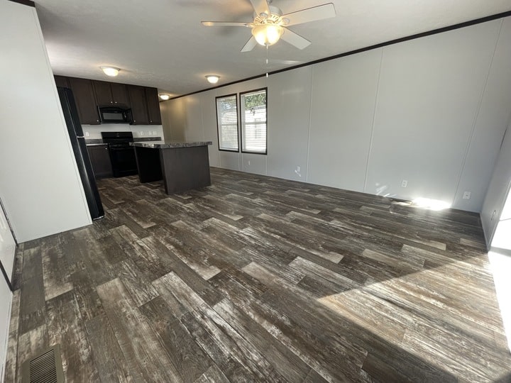 Open-concept living space with wood-patterned flooring, white walls, a ceiling fan, large windows, and a modern kitchen with dark cabinets, black appliances, and an island. Natural light fills the room.
