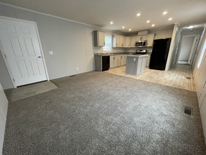 Open-plan living area with gray carpet, leading to a modern kitchen with light cabinets, stainless steel appliances, black refrigerator, and tile flooring. White front door and hallway are visible in the background.