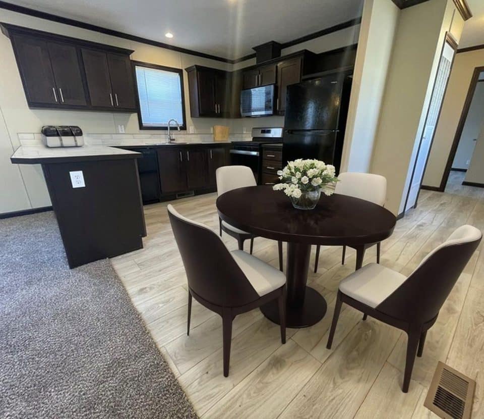 A modern kitchen and dining area with dark cabinets, a black fridge, and oven. A round dark table with four chairs and a floral centerpiece sits on light wood flooring next to carpeted living space.