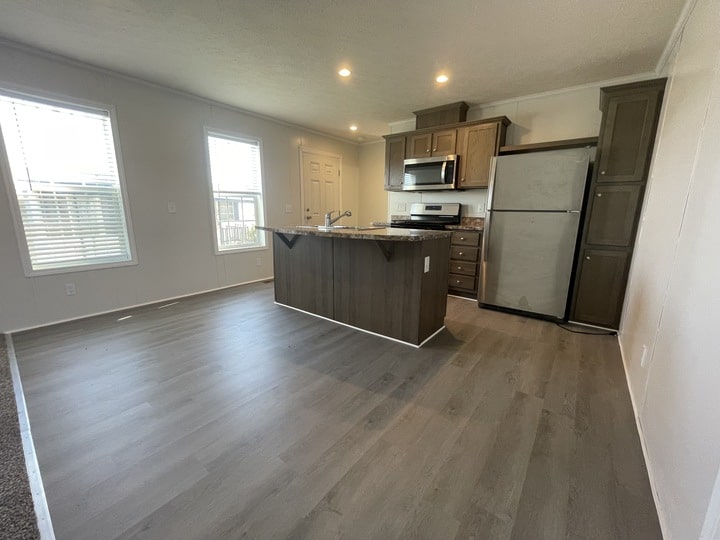 Modern kitchen with gray wood flooring, a central island with a sink, stainless steel appliances including a fridge and microwave, brown cabinets, and large windows letting in natural light.