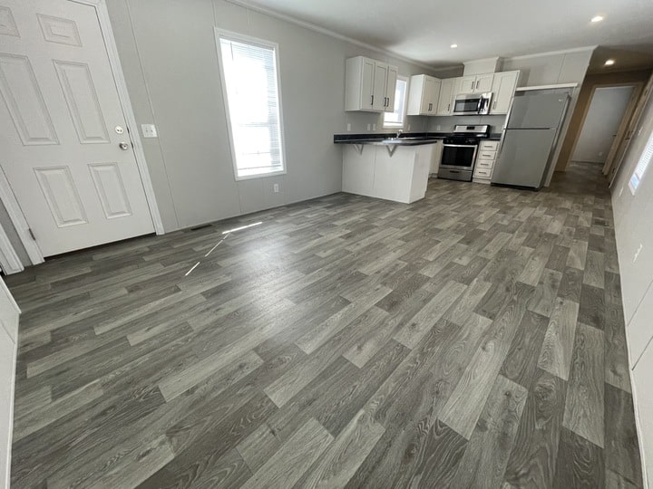 A modern kitchen and living area with gray wood-patterned flooring, white cabinets, stainless steel appliances, and large windows letting in natural light. The space is empty and has a clean, open layout.