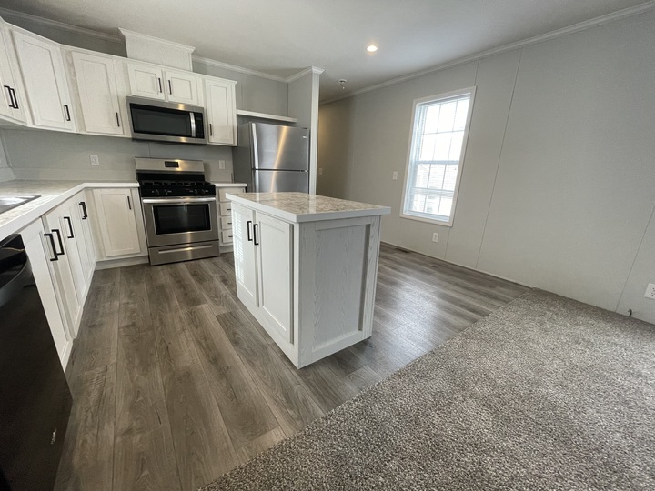 Modern kitchen with white cabinets, stainless steel appliances, an island with a marble-like countertop, and wood-look flooring. Adjacent is a carpeted living area and a large window letting in natural light.