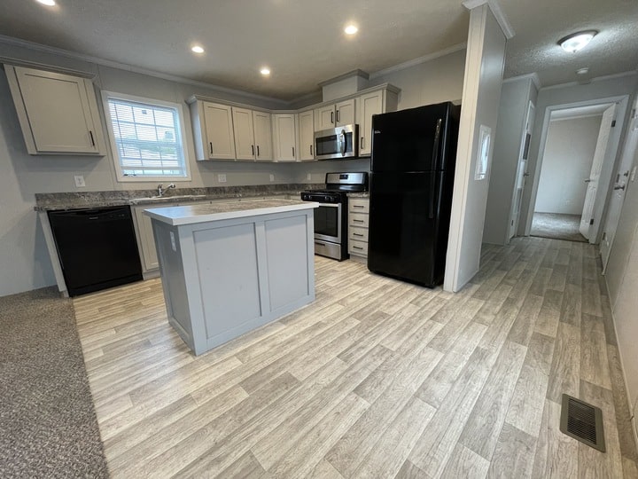 Modern kitchen with light gray cabinets, black appliances, an island, and wood-patterned vinyl flooring. Theres a window above the sink and a hallway leading to other rooms in the background.