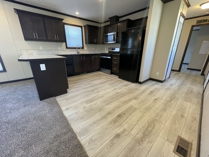 Modern kitchen with dark cabinets, black appliances, and light wood flooring next to a carpeted living area. There is a small island, white subway tile backsplash, and a window above the sink.