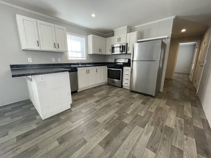 Modern kitchen with white cabinets, stainless steel appliances, black countertops, wood-style flooring, and a small island. A window above the sink lets in natural light, and a hallway is visible to the right.