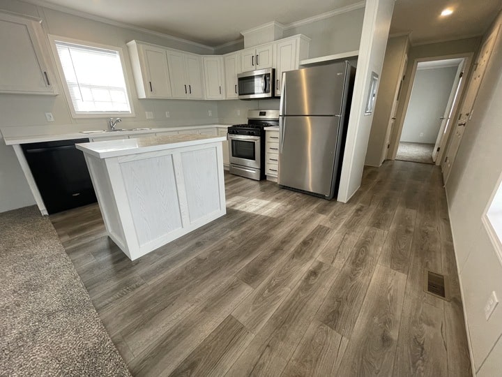 Modern kitchen with white cabinets, stainless steel appliances, and wood-look flooring. An island stands in the center, and a hallway leads to other rooms. Natural light enters through a window above the sink.
