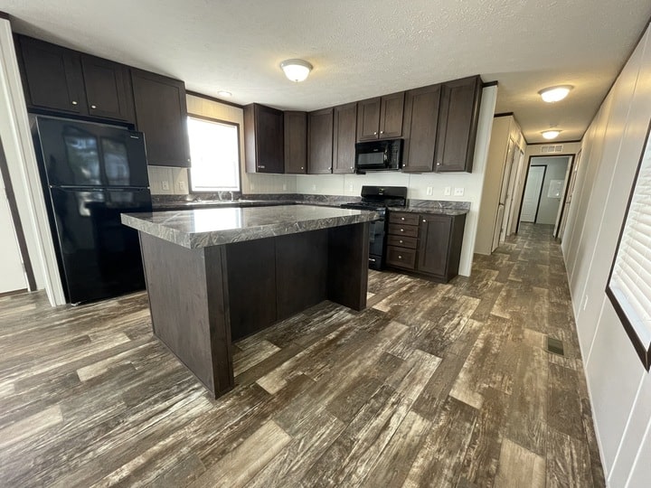 Modern kitchen with dark wood cabinets, a large island with marble-like countertop, black appliances, and wood-look flooring. A hallway extends to the right with additional rooms visible at the end.