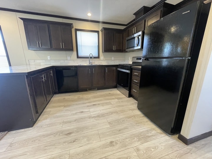 Modern kitchen with dark wooden cabinets, black appliances including a refrigerator, stove, dishwasher, and microwave, light-colored countertops, and light wood-look flooring. A window is above the sink.