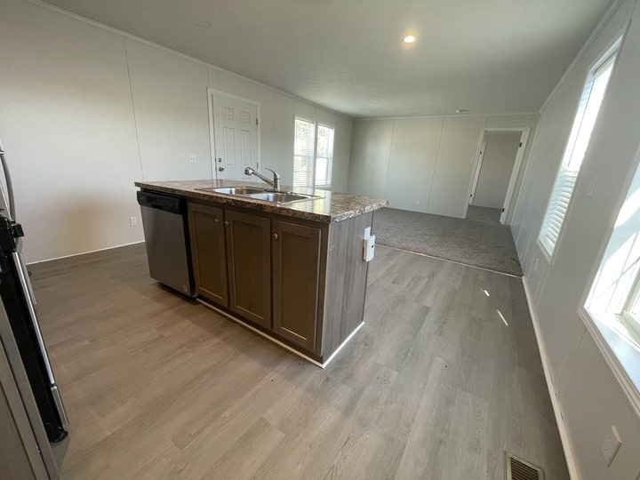A modern, unfurnished kitchen and living area with light wood floors, a center island with a sink and dishwasher, white walls, large windows, and a door leading to another room.