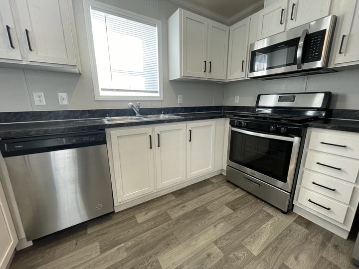 Modern kitchen with white cabinets, stainless steel appliances including a dishwasher, gas stove, and microwave, dark countertops, a double sink under a window, and wood-patterned vinyl flooring.