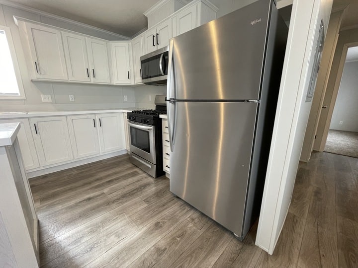Modern kitchen with white cabinets, stainless steel refrigerator and oven, microwave, light wood flooring, and neutral walls. A hallway with carpeted flooring is visible in the background.