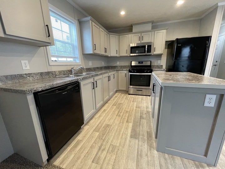 Modern kitchen with light gray cabinets, granite countertops, stainless steel stove and microwave, black refrigerator and dishwasher, wood-look flooring, and a window above the sink.