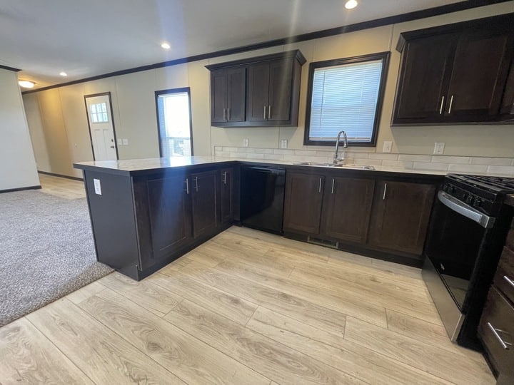 Modern kitchen with dark wood cabinets, light wood flooring, black appliances, a window above the sink, and an open layout leading to a carpeted living area and entryway.