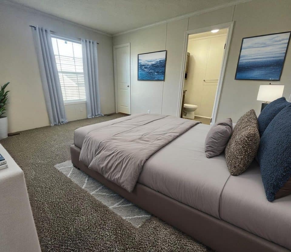 A modern bedroom with a gray bed, textured pillows, beige carpet, two ocean-themed paintings, a potted plant, and a door leading to a bathroom. Sunlight streams in through large windows with light curtains.