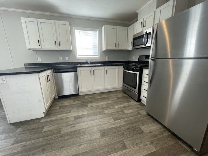Modern kitchen with white cabinets, black countertops, stainless steel appliances including refrigerator, oven, microwave, and dishwasher, light gray walls, and wood-look vinyl flooring. A small window above the sink lets in light.