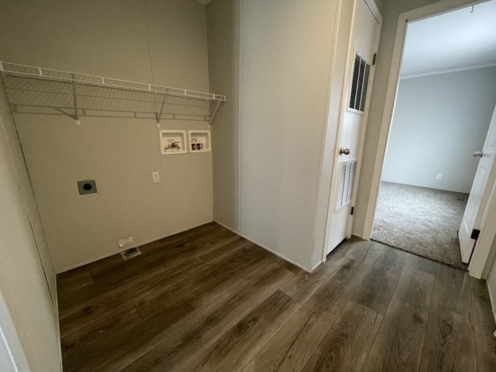 A small laundry room with wood-look flooring, a white wire shelf on the wall, two wall outlets, and a doorway leading to a carpeted room. Two small pictures are mounted on the wall beneath the shelf.