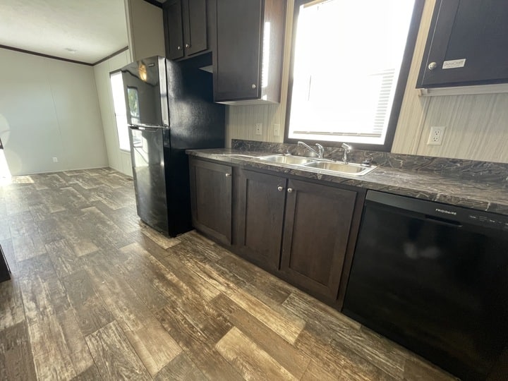 Modern kitchen with dark wood cabinets, black refrigerator, dishwasher, stainless steel sink, and marble-patterned countertops. Sunlight streams in through a window above the sink, illuminating the wood-look flooring.