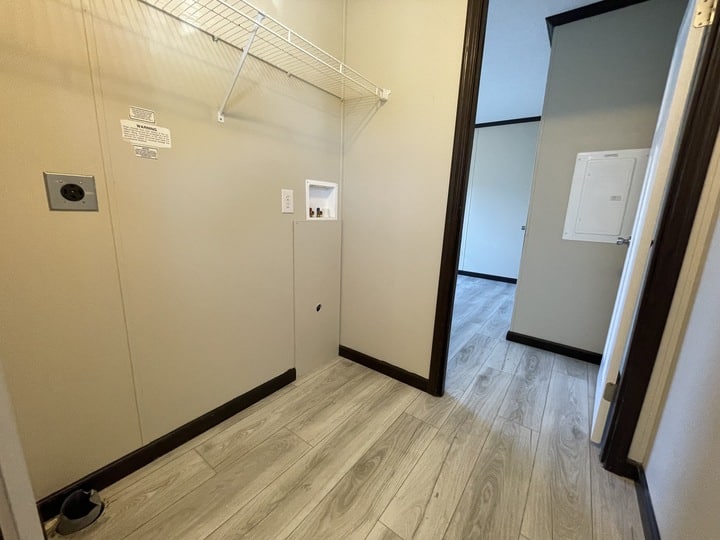 A laundry room with light wood flooring, beige walls, a wire shelf, washer and dryer hookups, and a utility panel. An open doorway leads to another room with similar flooring.