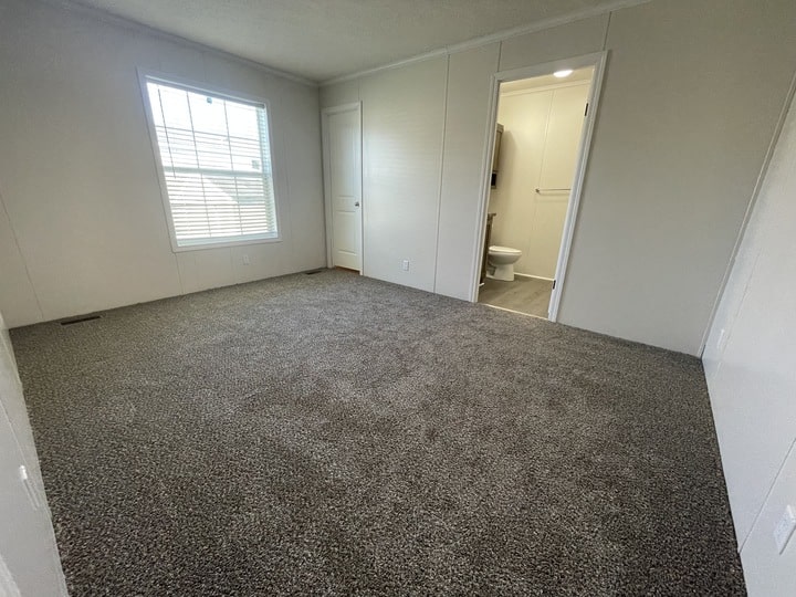 Empty bedroom with gray carpet, white walls, a large window on the left, and an open door leading to a bathroom with a visible toilet and towel rack.