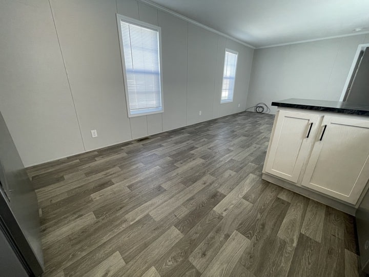 A room with light gray walls, wood-patterned vinyl flooring, two tall windows, and a partial view of a white kitchen cabinet with a dark countertop. The space is empty and well-lit by natural light.